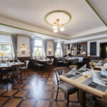 A bright, inviting restaurant dining room with wooden floors, neatly set tables with striped napkins, cushioned seating, large windows with curtains, and classic light fixtures on the ceiling.