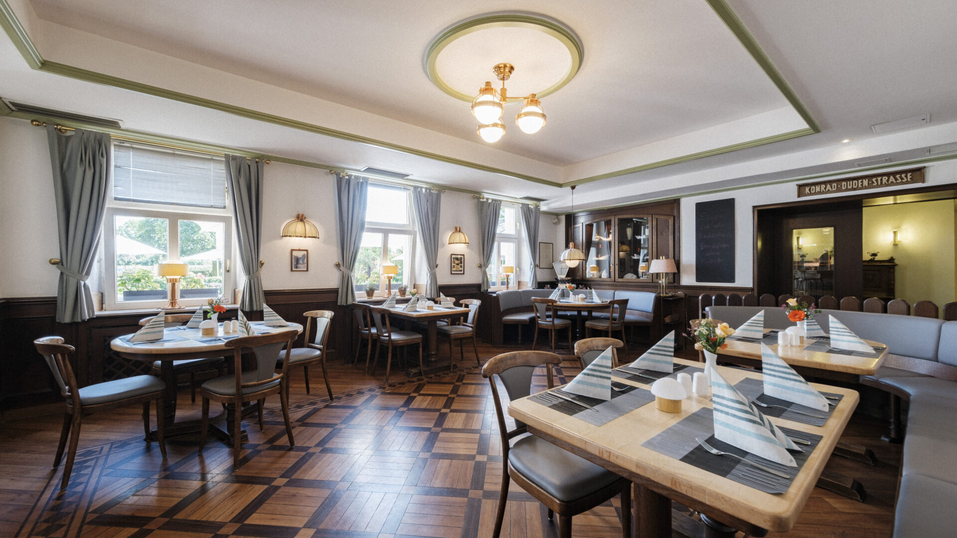 A bright, inviting restaurant dining room with wooden floors, neatly set tables with striped napkins, cushioned seating, large windows with curtains, and classic light fixtures on the ceiling.