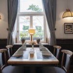 A neatly set dining table for four with striped napkins and a small flower vase, placed by a window with gray curtains in a cozy, softly lit restaurant. Two wall lamps and framed pictures decorate the walls.