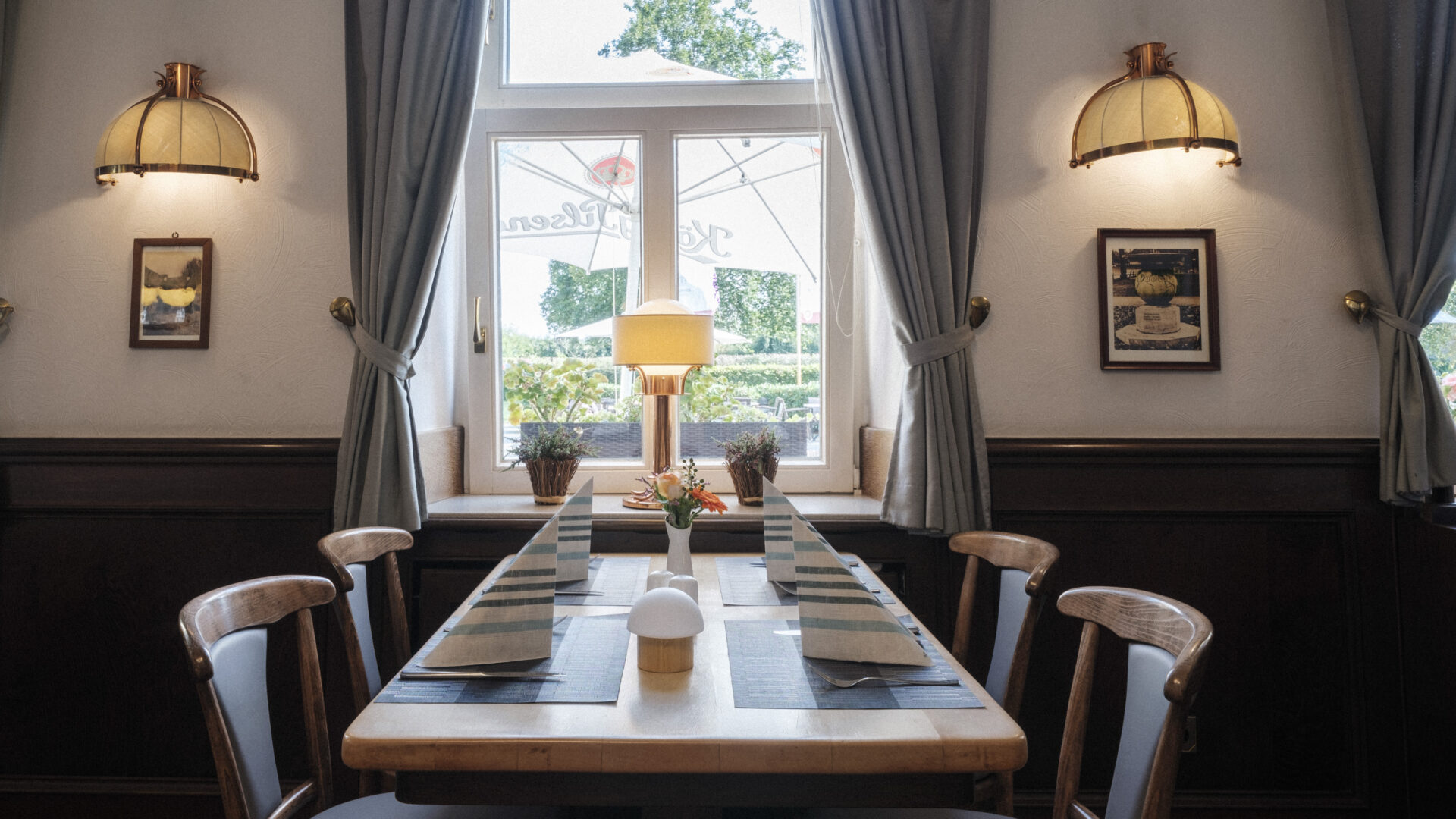 A neatly set dining table for four with striped napkins and a small flower vase, placed by a window with gray curtains in a cozy, softly lit restaurant. Two wall lamps and framed pictures decorate the walls.