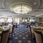 A spacious, elegant restaurant dining room with neatly arranged tables and chairs, blue patterned carpet, and a large decorative stained-glass ceiling light. Sunlight streams through large windows along the back wall.