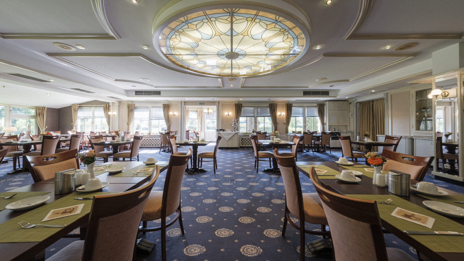 A spacious, elegant restaurant dining room with neatly arranged tables and chairs, blue patterned carpet, and a large decorative stained-glass ceiling light. Sunlight streams through large windows along the back wall.