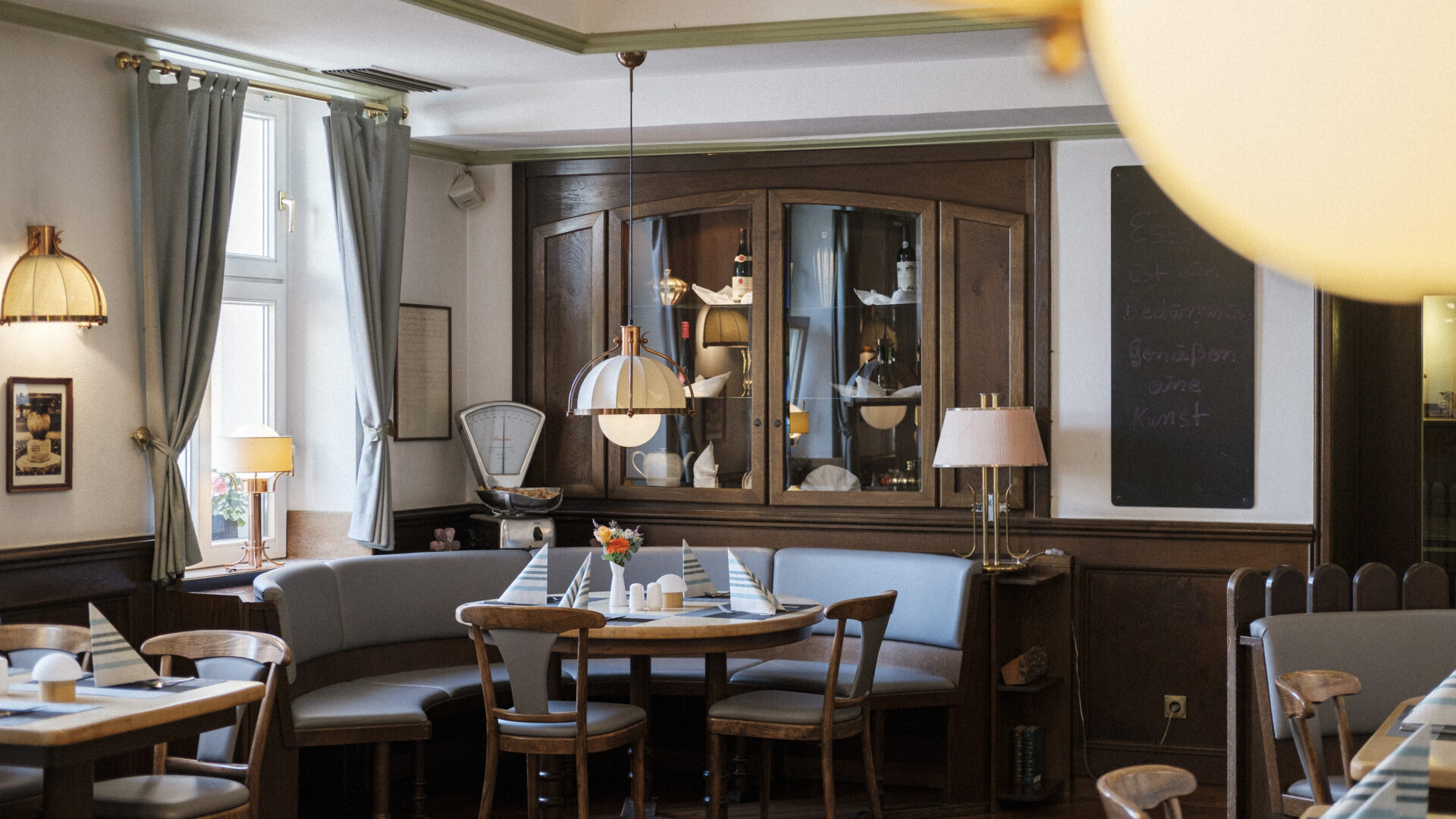 A cozy restaurant interior with wooden chairs and tables, blue upholstered benches, pendant lamps, and soft natural light from windows. The decor includes mirrors, framed art, a chalkboard, and fresh flowers on the tables.